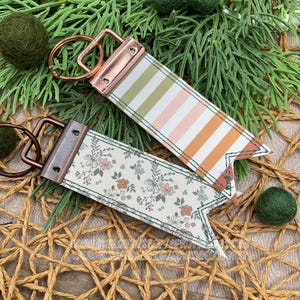 May include: Two keychains with copper-colored hardware. One has a striped pattern in shades of green, pink, and orange. The other features a floral design with pink and green flowers on a cream background. The keychains are on a woven surface.