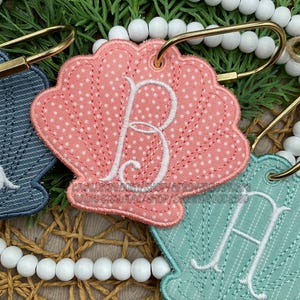 May include: A coral pink fabric seashell keychain featuring a white polka dot pattern and a white embroidered letter B. The keychain is attached to a gold-toned metal ring and is surrounded by white wooden beads and green pine needles.