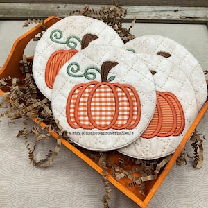 Puede incluir: Cuatro posavasos redondos de tela acolchada con diseños de calabazas bordadas en una bandeja de madera naranja. Las calabazas tienen patrones a cuadros o a rayas naranjas y blancos, vides verdes y tallos marrones. Los posavasos son de color crema con un patrón de diamantes.