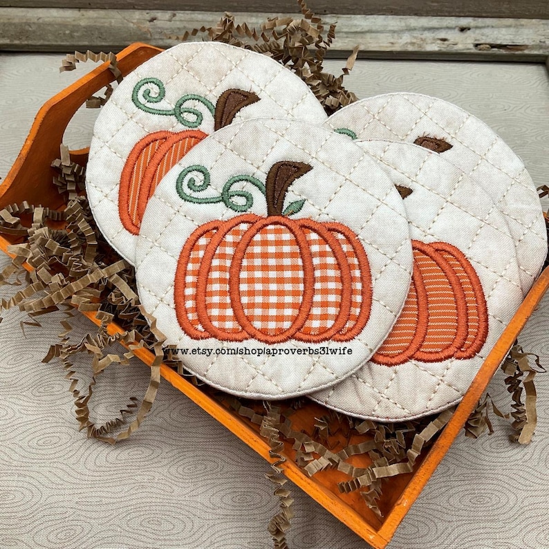 May include: Four round, quilted fabric coasters with embroidered pumpkin designs in an orange wooden tray. The pumpkins have orange and white gingham or striped patterns, green vines, and brown stems. The coasters are cream-colored with a diamond pattern.