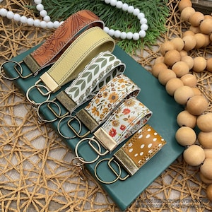 Puede incluir: Una colección de llaveros con varios diseños. Los llaveros presentan correas de cuero y tela en tonos marrón, amarillo y blanco, adornadas con patrones florales, de hojas y lunares. Cada llavero tiene un cierre metálico y un llavero.