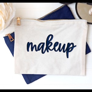 May include: A white canvas makeup bag with a zipper closure. The bag has the word "makeup" embroidered in dark blue on the front.