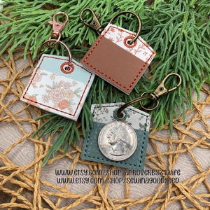 May include: Three square keychains with metal clasps. One keychain features a floral pattern, another has a brown leather section, and the third holds a quarter coin. The keychains are on a woven surface with green sprigs.
