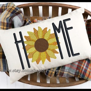 May include: A white pillow with the word "HOME" embroidered in black with a sunflower in the center. The pillow is sitting in a woven basket with a plaid blanket.