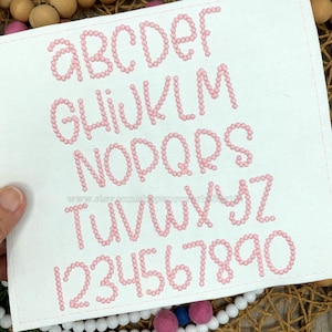 May include: A white fabric square with the alphabet and numbers embroidered in pink. The letters and numbers are made of small, connected circles. The text "www.etsy.com/shop/pieproeworks88" is visible.