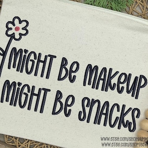 Puede incluir: Una bolsa de lona beige con cremallera con el texto "Might be makeup, might be snacks" en escritura negra. Una flor negra con un centro rojo está en la parte superior izquierda. La bolsa está sobre una superficie tejida con cuentas de madera.