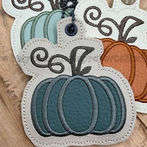 Può includere: Tre portachiavi in feltro a forma di zucca con un gambo marrone e un design a spirale. Le zucche sono turchese, arancioni e turchese scuro. I portachiavi sono attaccati a una catena argentata.