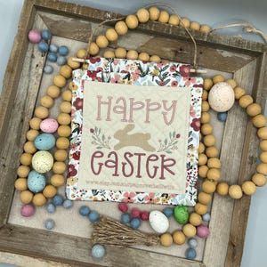 May include: A quilted fabric banner with a floral pattern and the text "Happy Easter" in pink and red lettering. A brown bunny silhouette is in the center of the banner. The banner is hanging from a wooden frame with a string of wooden beads and pastel colored eggs.