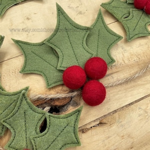 以下が含まれることがあります： 緑のフェルト製のホーリーの葉と赤いフェルト製のベリー。葉は緑色の糸で縫われており、ベリーは丸いです。葉とベリーは木製の表面に配置されています。