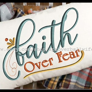 Puede incluir: Una almohada blanca con el texto "faith over fear" bordado en hilo turquesa, naranja y amarillo. La almohada está sobre una tela a cuadros.