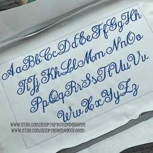 May include: A white fabric sheet featuring a full alphabet of uppercase and lowercase letters embroidered in blue thread using a cursive script font. The embroidery is framed by a thin blue rectangular border, with two URL links printed at the bottom.