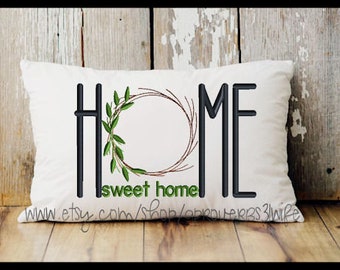 May include: White pillow with black embroidery that reads "HOME" with a green wreath in the center and the words "sweet home" below.