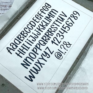 May include: White fabric with embroidered dark blue uppercase and lowercase letters, numbers, and symbols. The text includes the alphabet, numbers 1-9, and symbols. The fabric is held in a metal frame. The Etsy shop URLs are visible.