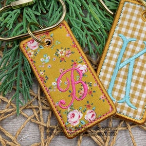 May include: A rectangular fabric keychain featuring a mustard yellow background with a floral pattern and a pink embroidered letter B. The edges are finished with a matching mustard yellow stitched border and a gold-toned metal split ring.