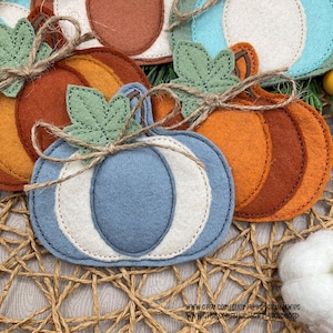 Puede incluir: Primer plano de varias decoraciones de calabaza de fieltro en tonos azules, naranjas y marrones. Cada calabaza tiene un diseño cosido y está adornada con una hoja de fieltro verde y un lazo de cuerda. Las calabazas están dispuestas sobre una superficie tejida.