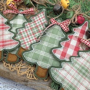 May include: Close-up of handmade Christmas tree ornaments. The ornaments are made of fabric with red and green plaid patterns, trimmed with green stitching and brown felt trunks. Each tree has a red and white checkered ribbon bow. The background includes bells and greenery.