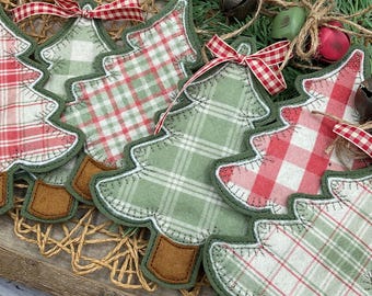May include: Close-up of handmade Christmas tree ornaments. The ornaments are made of fabric with red and green plaid patterns, trimmed with green stitching and brown felt trunks. Each tree has a red and white checkered ribbon bow. The background includes bells and greenery.