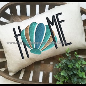 May include: Beige rectangular cushion embroidered with the word "HOME" in black capital letters. A seashell design in teal, brown, and gold is featured. The cushion is in a wooden basket with greenery.
