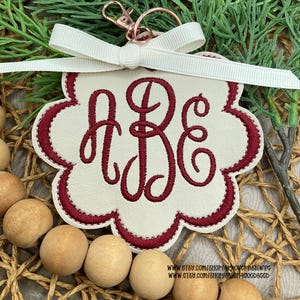 May include: A cream-colored keychain with a scalloped edge, featuring the embroidered letters "ABE" in a burgundy script. It has a burgundy border, a white ribbon, and a rose gold clasp. The background includes wooden beads and greenery.