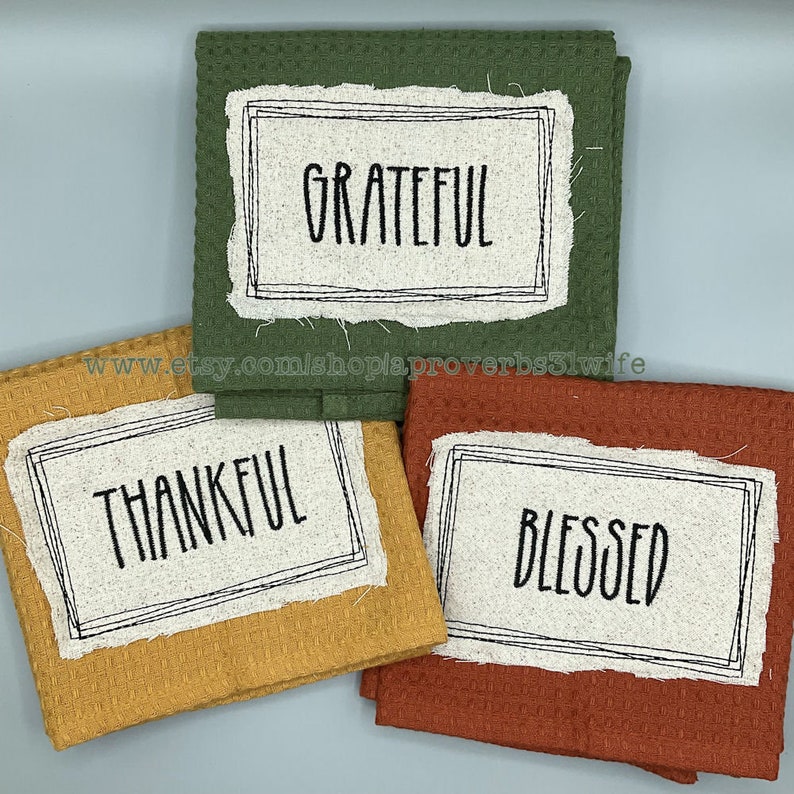 Puede incluir: Tres pa&ntilde;os de cocina de tejido de gofre en tonos verde, dorado y &oacute;xido. Cada pa&ntilde;o presenta un parche rectangular con un borde negro y una palabra en negro: "Grateful", "Thankful" y "Blessed".