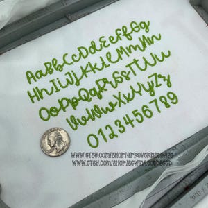 May include: A white fabric surface featuring green embroidered cursive letters of the alphabet in both uppercase and lowercase, along with the numbers 0 through 9. A silver United States quarter coin is placed next to the embroidered characters.