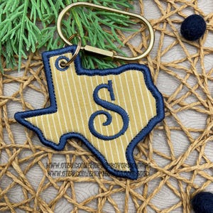 May include: A Texas-shaped keychain featuring a yellow fabric with thin white vertical stripes, embroidered with a dark blue letter S in the center. The keychain is outlined in dark blue thread and attached to a gold-toned metal oval ring.