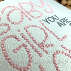 Puede incluir: Tela blanca con texto bordado en rosa y gris. El bordado rosa deletrea "GIRLS" y "BABES" en un estilo cursivo. El texto gris dice "YOU ARE". La tela tiene una apariencia texturizada.