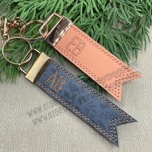 Puede incluir: Dos llaveros personalizados con herrajes dorados. Uno es de color melocotón con "EB" bordado, el otro es azul con "AB" bordado. Ambos tienen un borde cosido y un patrón floral.