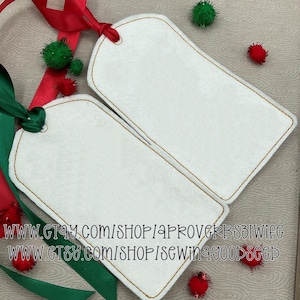 May include: Two white gift tags with brown stitched edges and a small hole at the top for ribbon. Red and green ribbons and small pom-poms are scattered around the tags. The text "WWW.ETSY.COM/SHOP/APROVERBS31WIFE" and "WWW.ETSY.COM/SHOP/SEWIN4GOODSEED" is visible.