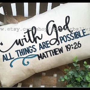 Puede incluir: Cojín rectangular color crema con texto bordado: "with God, all things are possible... Mateo 19:26". El texto está en escritura negra y azul. El cojín está sobre una cesta tejida.