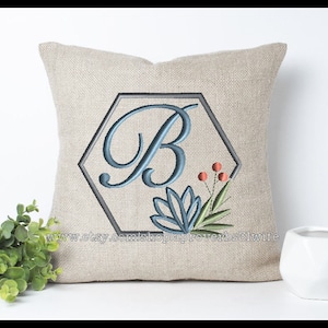 May include: A beige cushion with an embroidered design. The design features a blue letter "B" inside a grey hexagon, with floral accents. The cushion is on a white surface.