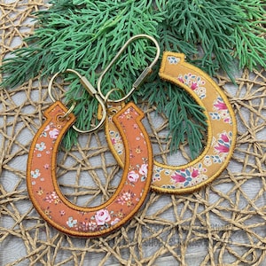 May include: Two horseshoe-shaped fabric keychains with metal lobster clasps. One keychain features a burnt orange fabric with a white and pink floral pattern, while the other is mustard yellow with a pink, blue, and white floral print.