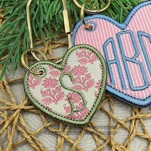 May include: Two heart-shaped fabric keychains with gold-tone metal rings. One keychain features a cream background with a pink floral pattern and a green embroidered letter S. The other has pink and white stripes with a blue embroidered monogram.