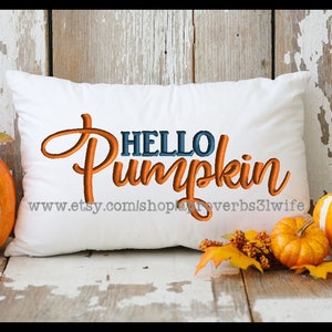 May include: A white pillow with the text "HELLO PUMPKIN" embroidered in orange and blue. The pillow is sitting on a wooden surface with two pumpkins and fall foliage.