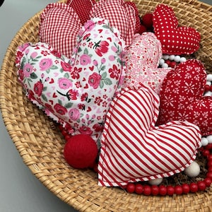 Puede incluir: Una cesta de mimbre llena de almohadas con forma de corazón rojas y blancas. Las almohadas están hechas de diferentes telas, incluyendo vichy, estampado floral y rayas.