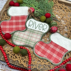 May include: Three Christmas stocking ornaments with red and green plaid patterns. One stocking has the name "RIVER" embroidered on the cuff. The stockings are decorated with red and green felt balls, bells, and greenery, arranged on a woven tray.