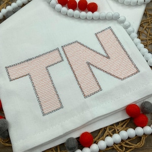 Puede incluir: Tela blanca doblada con las letras "TN" bordadas en coral con un borde gris. La tela está decorada con cuentas rojas, grises y blancas. Las letras miden aproximadamente 10 cm de alto.