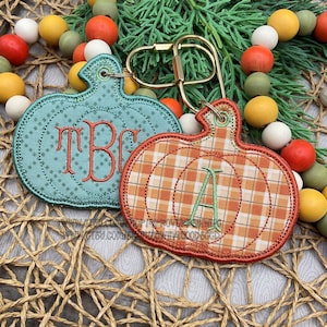 May include: Two embroidered pumpkin-shaped keychains with gold-tone metal hardware. One is teal with a diamond pattern and the orange monogram TBC, while the other is orange and white plaid with a green embroidered letter A.