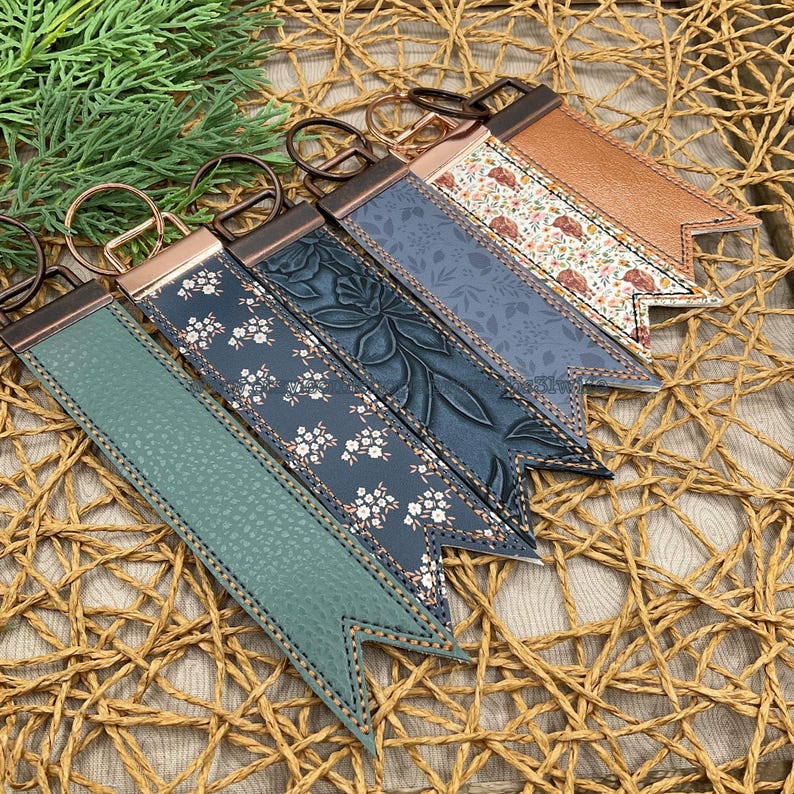 May include: A collection of keychains with various designs. The keychains feature a leather strap with different patterns, including floral and textured designs, and a metal ring. The color palette includes shades of blue, green, and brown.