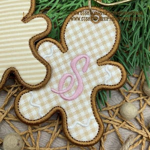 May include: A gingerbread man-shaped ornament featuring a beige and white houndstooth pattern, a stitched pink letter S in the center, and brown embroidered edges. The ornament includes a metal ring for hanging.