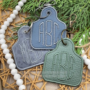 May include: Three leather keychains in shades of blue and green, each featuring embroidered initials. The keychains have metal hardware and are arranged on a woven surface with decorative elements.