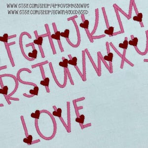 May include: Close-up of embroidered pink letters of the alphabet and the word "LOVE" on a light blue fabric. Each letter is adorned with small red heart-shaped embellishments. The image is a close-up of the embroidery.