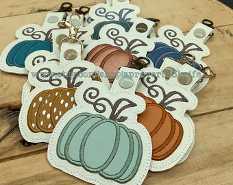 May include: A collection of pumpkin-shaped keychains in various colors, including teal, orange, and brown, with white accents and gold-colored clasps. Each keychain features a snap closure and a decorative vine design.