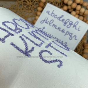 May include: Close-up of white fabric with embroidered lavender text. The word "SOUL" is prominently displayed. A small card with the alphabet in lavender script is in the background. The Etsy shop URL is visible at the bottom.