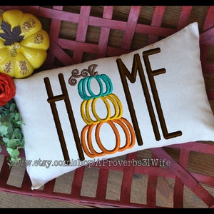 Puede incluir: Una almohada blanca con la palabra "HOME" bordada en marrón, con una pila de tres calabazas en el centro de la palabra. Las calabazas son naranja, amarilla y turquesa. La almohada está sentada en una cesta de mimbre roja.