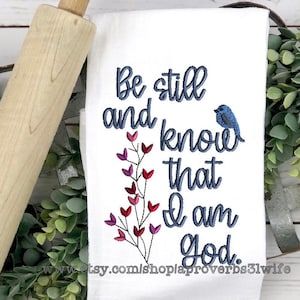 以下が含まれることがあります： 白いキッチンクロスに、青い刺繍で「静かにしなさい。私は神であることを知りなさい。」と書いてあります。タオルの右側には小さな青い鳥が刺繍されています。タオルの左側にはピンクと赤の刺繍の花があります。