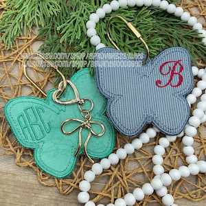 May include: Two butterfly-shaped keychains. One is teal with an embroidered monogram, the other is denim blue with a red letter "B". Both have gold-colored hardware and are displayed with white beads and greenery.
