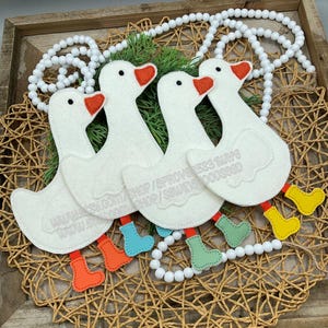 May include: Four white felt goose ornaments with orange beaks and feet in various colours. The geese are arranged on a woven tray with white bead garland and greenery. Text is visible on the geese.
