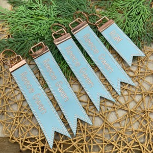 May include: A collection of personalized keychains. Each keychain features a light blue ribbon with the name "Reece Gwen" or initials "RGM" embroidered in a copper color. The keychains have copper-colored metal hardware and are arranged on a woven surface with greenery.