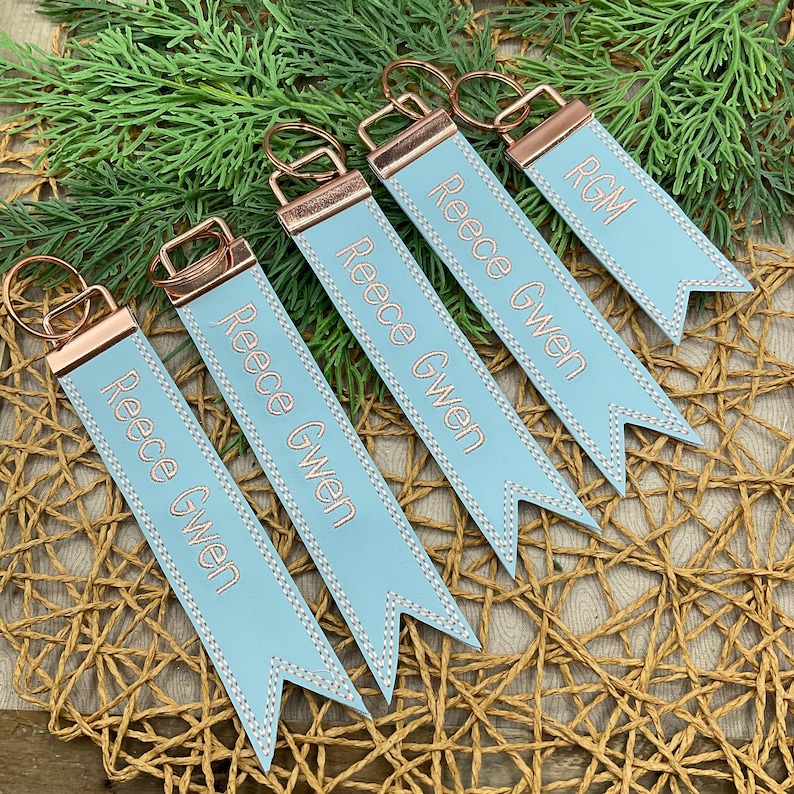 May include: A collection of personalized keychains. Each keychain features a light blue ribbon with the name "Reece Gwen" or initials "RGM" embroidered in a copper color. The keychains have copper-colored metal hardware and are arranged on a woven surface with greenery.